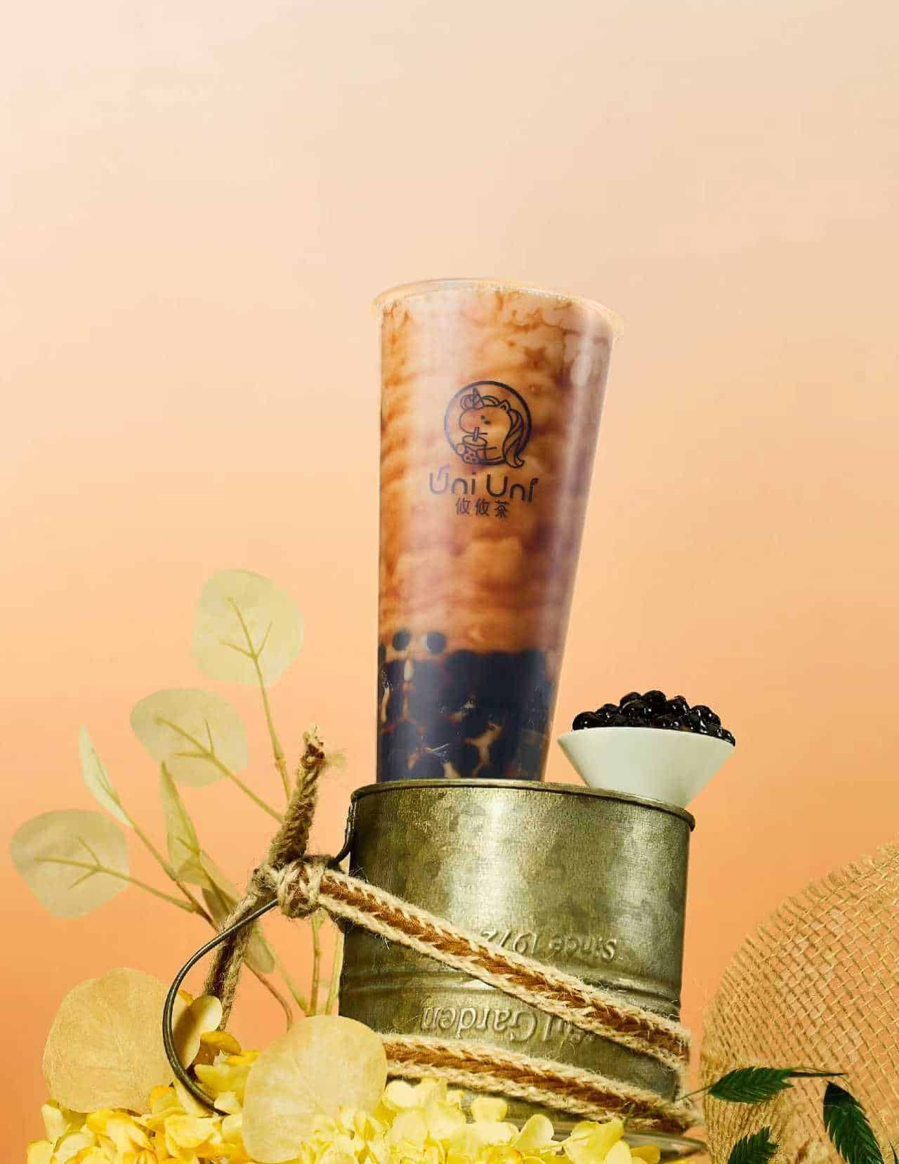 A tall cup of bubble tea with tapioca pearls, placed on a metal container, next to a small bowl of pearls, dried flowers, and a sun hat against a peach background. Uni Uni About Us. A tall cup of bubble tea with tapioca pearls, placed on a metal container, next to a small bowl of pearls, dried flowers, and a sun hat against a peach background.