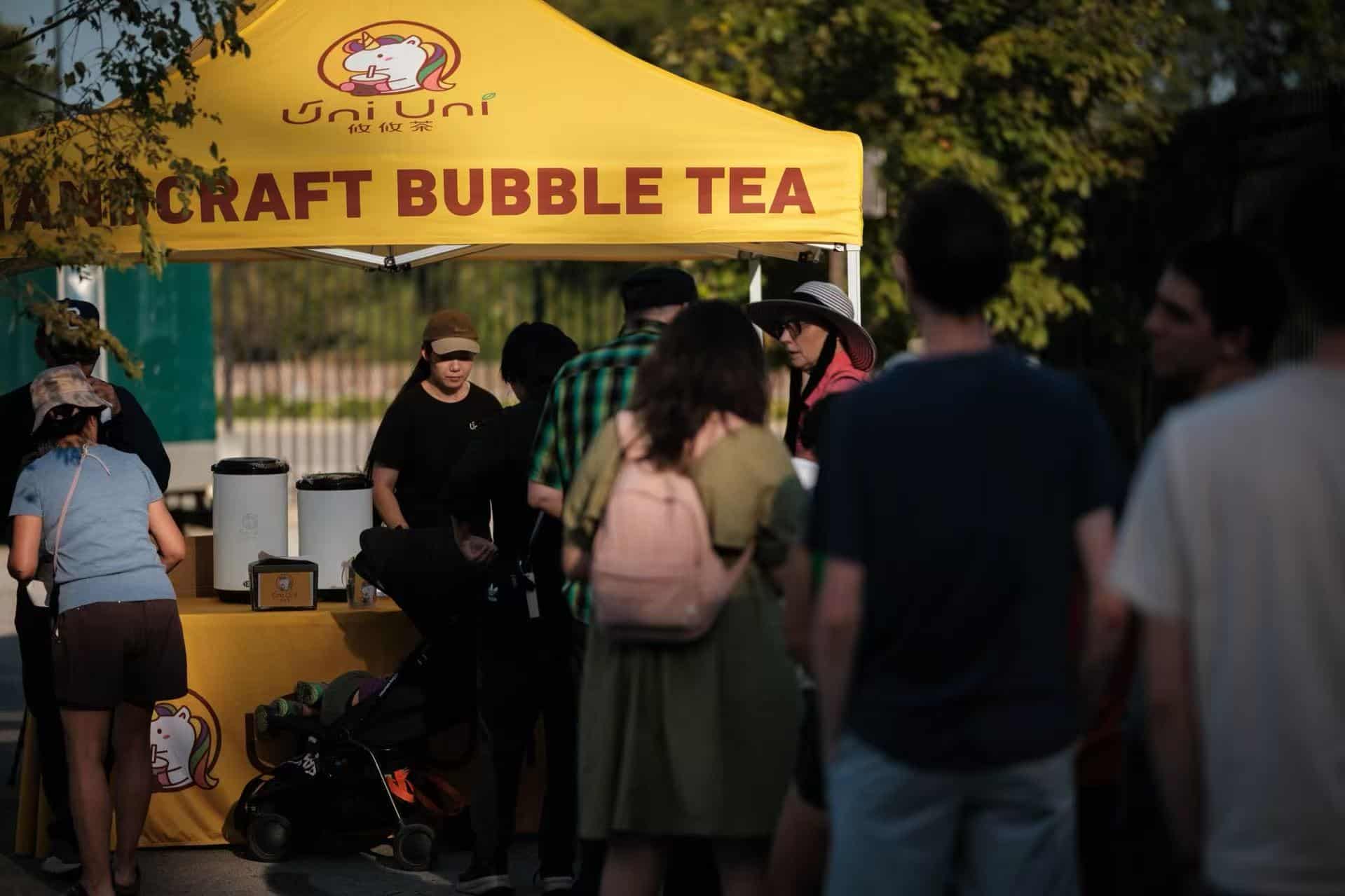 Uni Uni Boba Catering: Community and Schools. People gather in front of a yellow tent labeled "Handcraft Bubble Tea" with staff preparing drinks at an outdoor event.