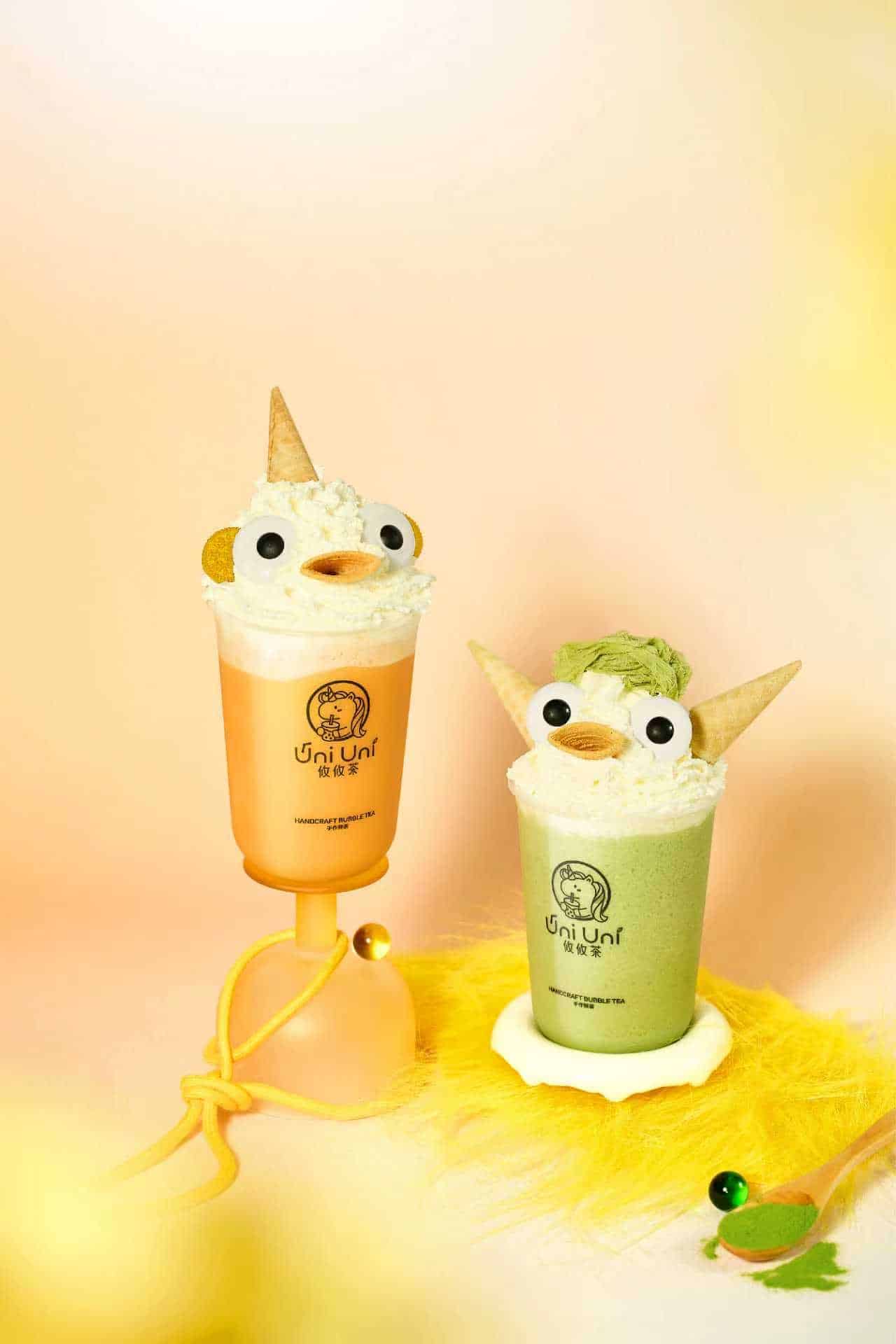 Two whimsical drinks with whipped cream, cookie eyes, and cone ears are served in cups, one orange and one green, against a light yellow background. Two whimsical drinks with whipped cream, cookie eyes, and cone ears are served in cups, one orange and one green, against a light yellow background. Creative Influencers.