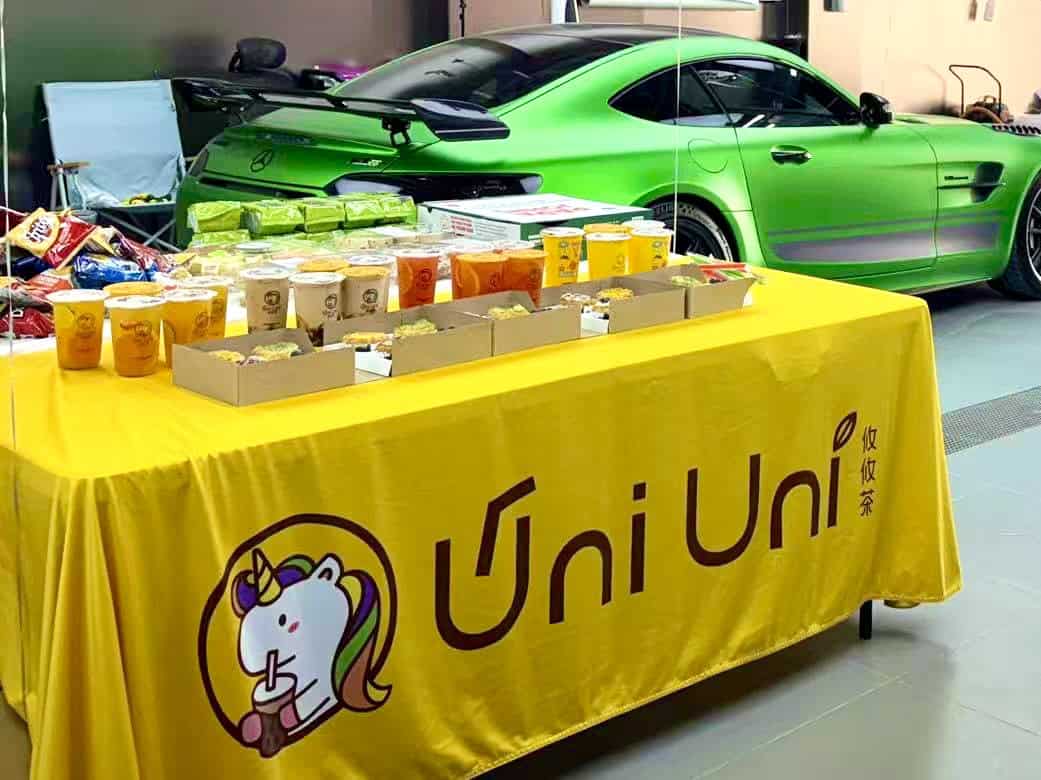 Uni Uni Catering Parties. A table with a yellow Uni Uni tablecloth displays packaged food and drinks in front of a green sports car in a garage.